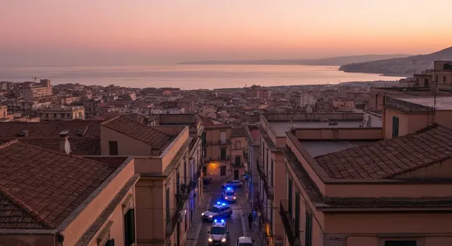 Blitz Ndrangheta Reggio Calabria: auto della polizia con lampeggianti blu all'alba tra i tetti della citta sullo Stretto